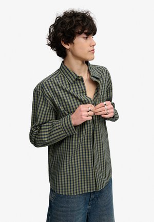 Junge Person mit lockigem Haar knöpft ein grün-navy kariertes Hemd zu, trägt mehrere Ringe und Jeans, vor einem schlichten Hintergrund.