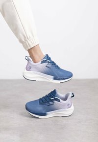 Zapatillas deportivas en azul y morado con parte superior texturizada, cordones prominentes y suelas blancas acolchadas. Incluyen un logo en el costado y una pestaña en el talón.