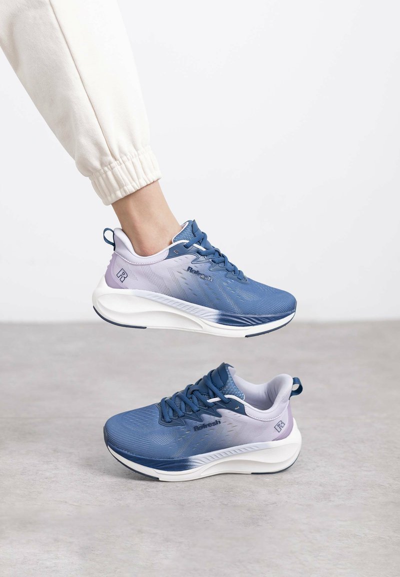 Zapatillas deportivas en azul y morado con parte superior texturizada, cordones prominentes y suelas blancas acolchadas. Incluyen un logo en el costado y una pestaña en el talón.