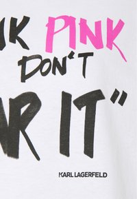 T-shirt bianco in cotone con testo in grassetto nero e rosa: "PENSA IN ROSA, NON INDOSSARLO." Include il nome "KARL LAGERFELD" in nero.