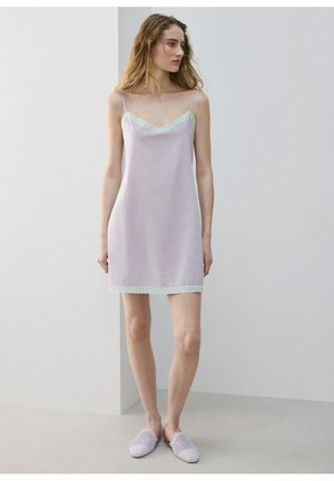 Tânără purtând o rochie subțire mov deschis cu bordură din dantelă și pantofi slip-on tricotati mov deschis, stând în fața unui perete simplu.