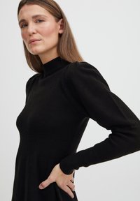 Melnā adīta kleita ar augstu apkakli, pieguļošu korpusu un pūkainām piedurknēm. Ar ribotu tekstūru un plūstošu svārku.