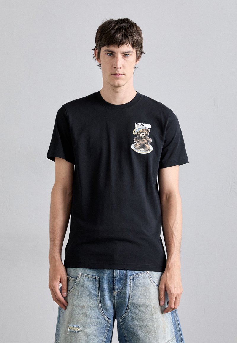 Svart bomull t-skjorte med grafisk design av en bjørn og teksten "Moschino" på venstre bryst. Modellen har på seg lyseblå, slitte jeans.