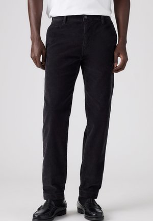 Homme portant un pantalon en velours côtelé noir, des chaussures noires et une chemise blanche, présenté de la taille aux pieds sur un fond uni.
