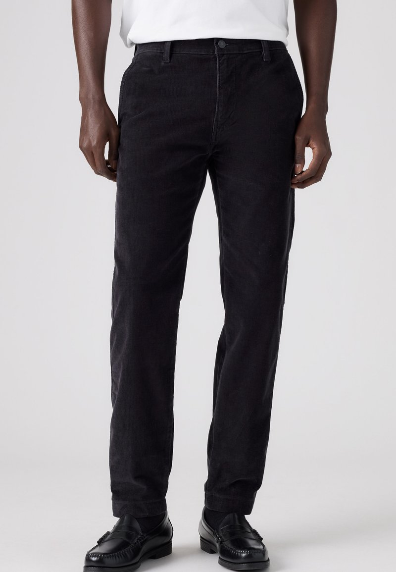 Homme portant un pantalon en velours côtelé noir, des chaussures noires et une chemise blanche, présenté de la taille aux pieds sur un fond uni.