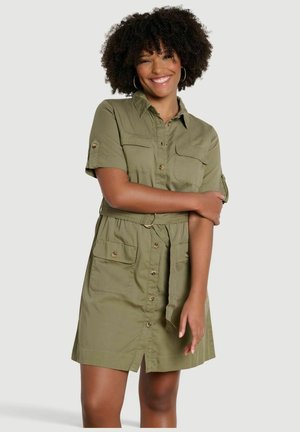 Uśmiechnięta kobieta o kręconych włosach ma na sobie oliwkową sukienkę z przodu zapinaną na guziki do kolan, z krótkimi rękawami, paskiem i dużymi przednimi kieszeniami.