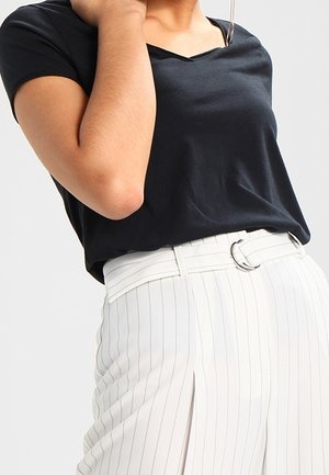 Person trägt ein schwarzes kurzärmeliges Oberteil, das in weiße, mit feinen Streifen versehene Hosen mit silberner Schnallengürtel gesteckt ist.