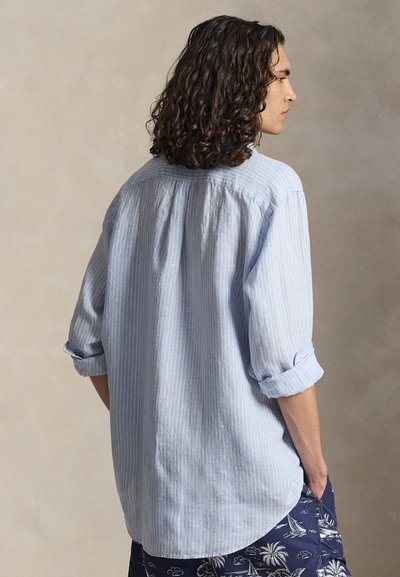 Jong persoon met schouderlang krullend haar, draagt een lichtblauw gestreept overhemd met opgerolde mouwen en marineblauwe korte broek met witte tropische patronen.