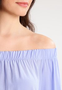 Blouse dégageant les épaules en tissu lavande clair avec un élastique au niveau du col et une texture douce et fluide. Design simple et fonctionnel.