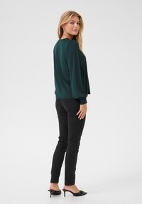 Blouse vert foncé à manches bouffantes, associée à un jean skinny noir et des mules à talons noires. Texture de tissu lisse, design simple.