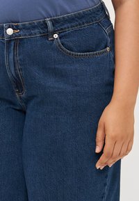 Zapleten prikaz roke osebe, ki počiva ob temno modrih kavbojkah z vidnimi šivi in podrobnostmi sprednjega žepa.