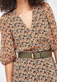Vestido floral con mangas abullonadas en crema, naranja y verde sobre un fondo negro, ceñido a la cintura con un cinturón verde que tiene una hebilla dorada.