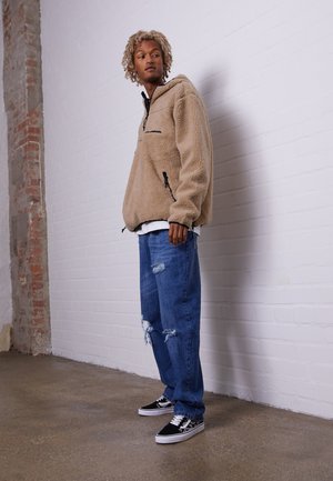Beige fleece sweatshirts med halv-lynlås, lange ærmer og sorte accenter. Et par slidte blå jeans og sneakers med sort-hvidt mønster.