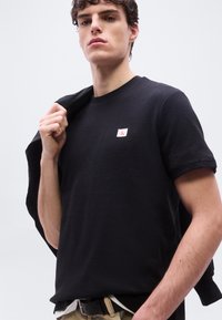 Νεαρός άνδρας που φοράει μαύρο υφασμάτινο t-shirt με μικρό λογότυπο CK στο στήθος, κρατώντας ένα μαύρο σακάκι στον ώμο του.