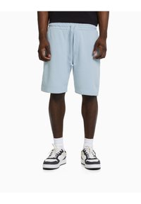 Muž v světle modrých šortkách na stažení, bílých ponožkách a černo-bílých teniskách, stojící na bílém pozadí.