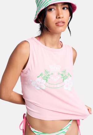 Jeune femme portant un haut sans manches rose avec un blason floral et un chapeau bob rayé rose-vert, levant l’ourlet pour montrer un bas de bikini rayé.