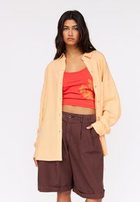 Světlé oranžové oversized tričko, červený top s květinovou výšivkou a hnědé skládání šortky. Měkké materiály, uvolněný střih a detaily s knoflíky.
