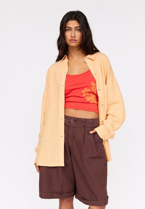 Light orange oversized shirt, red tank top with flower embroidery, and brown pleated shorts. Soft textures, relaxed fit, and button details.