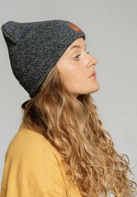 Szara dziana czapka z teksturowanym wykończeniem i małą skórzaną łatką. Kręcone, jasnobrązowe włosy częściowo widoczne, nosi żółtą koszulę.