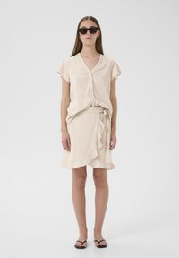 Gai beige linu apautēts svārki ar viļņotu apakšu, apvienots ar atbilstošu pogu augšu. Modelis nēsā melnas saulesbrilles un vienkāršas melnas sandales.