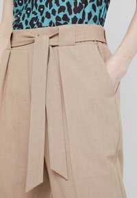 Beige hoog-geknipte broek met een gestrikte riem, plooien aan de voorkant en zijzakken, met een textuur stof en strakke lijnen.