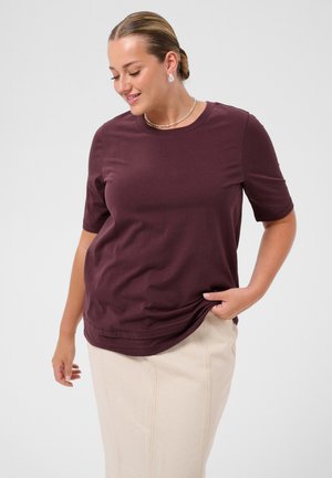 Vrouw met haar in een knot, pareloorbellen, een bordeauxrood T-shirt met korte mouwen en een beige rok, glimlachend en naar beneden kijkend met hand op de zoom van het T-shirt.