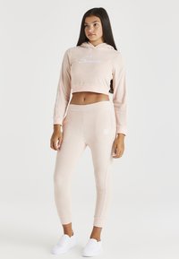 Corta sudadera rosa con logo en blanco, combinada con pantalones de chándal ajustados a juego que presentan detalles laterales y cintura elástica. Textura suave.