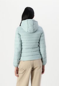 Lys blå puffjakke med hette, med kviltet tekstur og elastiske mansjetter. Båret med beige cargobukser, vist fra baksiden.