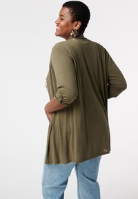 Cardigan long olive en vert avec des manches trois-quarts, texture douce et fentes latérales, associé à un jean léger bleu clair ample.