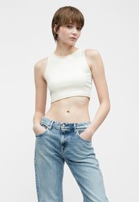 Persoană tânără cu părul scurt castaniu, purtând un top alb fără mâneci și blugi deschiși la culoare, cu talie înaltă, având mâinile în buzunare.