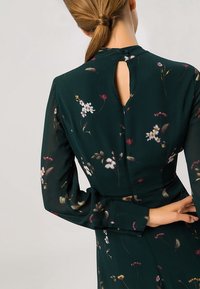 Kvinna i mörkgrön blommig klänning med långa ärmar, nyckelhål baktill och knappstängning, med händerna korsade bakom ryggen.