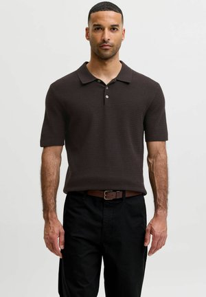 Uomo con i capelli corti che indossa una polo marrone scuro a maniche corte e pantaloni neri con cintura marrone, in piedi davanti a uno sfondo chiaro e uniforme.