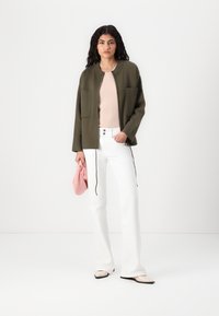 Olijfgroene jas met twee voorzakken, gecombineerd met een lichtroze top en witte wijde broek; een roze tas vasthoudend, met beige platte schoenen.
