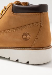 Botines de ante marrón con un collarín acolchado marrón, suela de goma blanca y logotipo de árbol grabado en el costado, con detalles de costura en contraste.