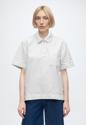 Jong persoon met kort blond haar, draagt een los wit poloshirt met korte mouwen en blauwe spijkerbroek, staand tegen een effen lichte achtergrond.
