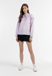 Lichtpaarse blouse met gestructureerde stippen, ruches bij de kraag en lange mouwen; gecombineerd met zwarte denim shorts en witte sneakers.