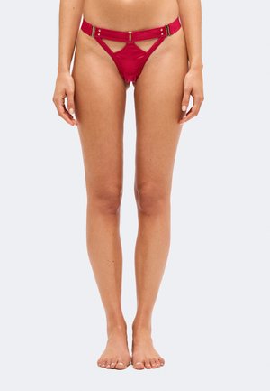 Femme portant un bas de bikini rouge à bretelles, debout pieds nus sur un fond clair uni.