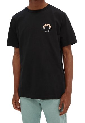 Mann trägt ein schwarzes T-Shirt mit kurzen Ärmeln und kleinem beigen und weißen "Scotch & Soda"-Logo auf der oberen linken Brust sowie hellblaue Jeans.
