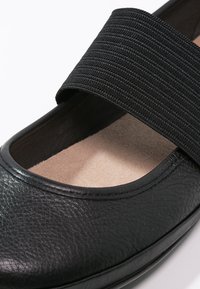 Zapato de cuero negro con una tira elástica texturizada atravesando el mediopié y una plantilla de color claro. Punta redondeada y acabado de superficie suave.