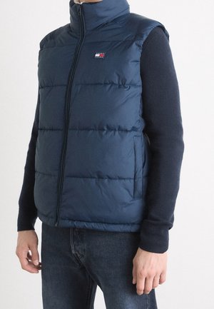 Homme portant une veste matelassée bleu marine sur une chemise noire à manches longues et un jean foncé, debout devant un fond blanc uni.