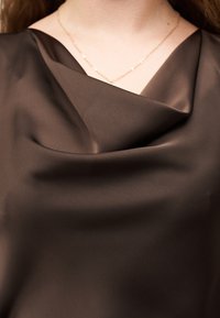 Κοντινό πλάνο ενός ατόμου που φοράει σκούρο καφέ σατέν τοπ με ντραπέ ντεκολτέ και λεπτή χρυσή αλυσίδα.