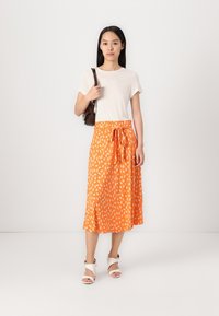 Oranger Midirock mit weißem Blättermuster, an der Taille gebunden. Kombiniert mit einem schlichten cremefarbenen T-Shirt und weißen Absatzsandalen, dazu eine braune Tasche.