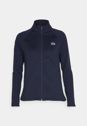 Chaqueta deportiva de color marino con cuello alzado, cremallera completa y mangas largas. Presenta un pequeño logo de cocodrilo en verde y blanco en el lado izquierdo del pecho.