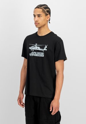 Joven con cabello trenzado lleva una camiseta negra con un helicóptero blanco y el texto "Alpha Industries Est. October 1959" en el frente.