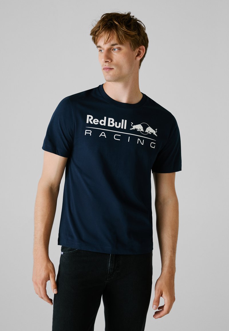 Junger Mann mit hellbraunen Haaren trägt ein dunkelblaues Red Bull Racing T-Shirt und eine schwarze Hose, blickt nach links vor einem grauen Hintergrund.