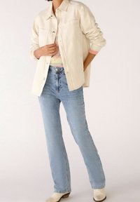 Giacca button-up beige chiaro con due tasche sul petto, abbinata a jeans a zampa d'elefante blu e stivali alla caviglia off-white.