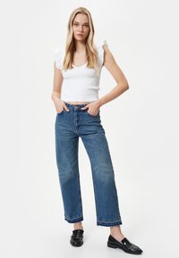 Weißes, geripptes Cropped-Top mit rüschierten Ärmeln, kombiniert mit blauen Schlagjeans mit ausgefransten Säumen und schwarzen Loafers.