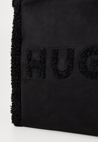 Czarna torba z teksturowanej tkaniny z puszystym wykończeniem. Na przedniej części znajduje się wytłoczone słowo "HUG" o kontrastowej, aksamitnej fakturze.