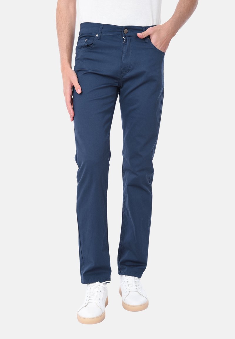 Pantaloni in cotone blu navy con vestibilità slim, dotati di tasche laterali e chiusura con bottone, abbinati a sneakers bianche e una camicia bianca semplice.