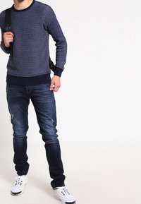 Pull en maille texturée marine avec des poignets et un ourlet côtelés, associé à un jean skinny bleu foncé et des baskets blanches. Sac à dos porté sur l'épaule gauche.